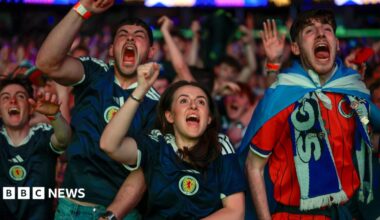 Fans celebrate in the OVO Hydro as Scotland score against Haiti. In the foreground are three men and one woman, all cheering and wearing Scotland strips. Rows of supporters can be seen behind them.