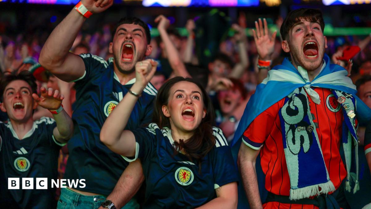 Fans celebrate in the OVO Hydro as Scotland score against Haiti. In the foreground are three men and one woman, all cheering and wearing Scotland strips. Rows of supporters can be seen behind them.