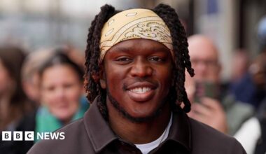 KSI smiles and wears a light yellow bandana over his hair with a white t-shirt and black shirt on top.