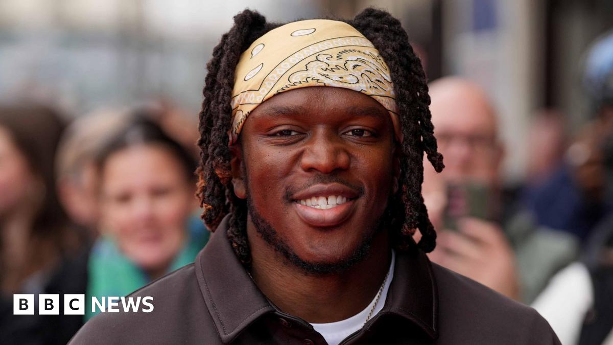 KSI smiles and wears a light yellow bandana over his hair with a white t-shirt and black shirt on top.