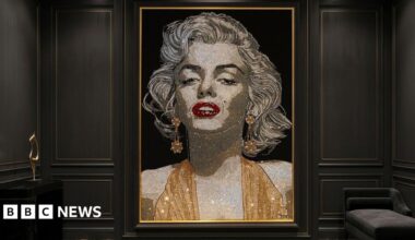 Alexandra is beaming at the camera. She has shoulder length brown hair and is is wearing a navy blue or black shirt. She appears to be standing inside at one of her exhibitions, with her artwork on a wall behind her.