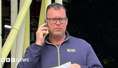 David Stroud, 44, after pleading guilty to harassing a woman on a train because of her sex. He is wearing a blue zip-up top, glasses, jeans and is holding a letter in one hand and a phone to his ear with the other.