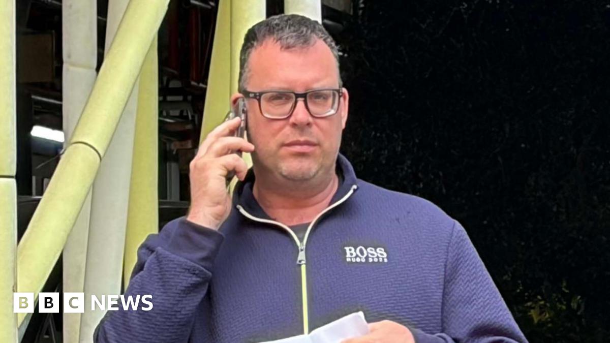 David Stroud, 44, after pleading guilty to harassing a woman on a train because of her sex. He is wearing a blue zip-up top, glasses, jeans and is holding a letter in one hand and a phone to his ear with the other.