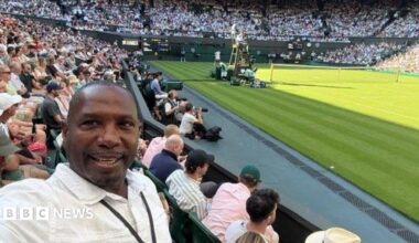 A man in a white collared shirt with a black lanyard around his neck is taking a selfie. He is sitting inside a tennis court, with a rows of audiences in the background.