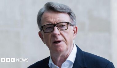 Lord Mandelson wears glasses, a white shirt and blue suit jacket, he is standing outside a stone building