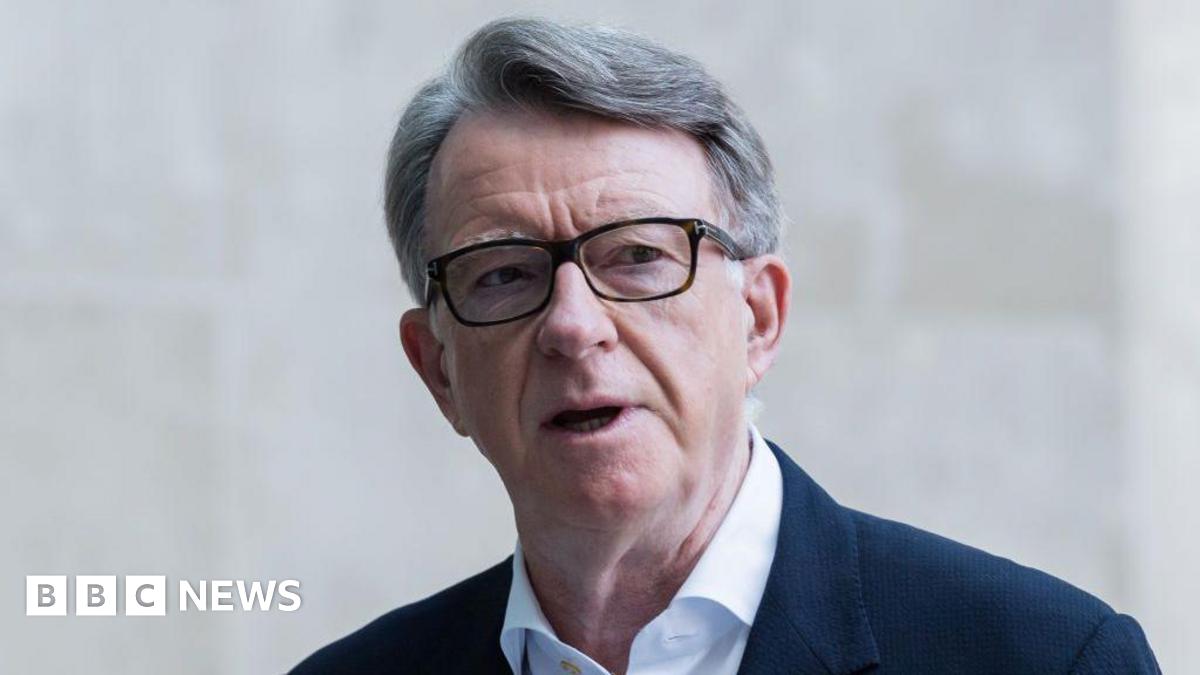 Lord Mandelson wears glasses, a white shirt and blue suit jacket, he is standing outside a stone building