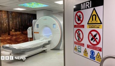 MRI scanner in a hospital room, which has an open door with a warning sign on saying MRI Strong Magnetic Field ALWAYS ON! The machine has a long flat bed that goes into a doughnut like scanner.