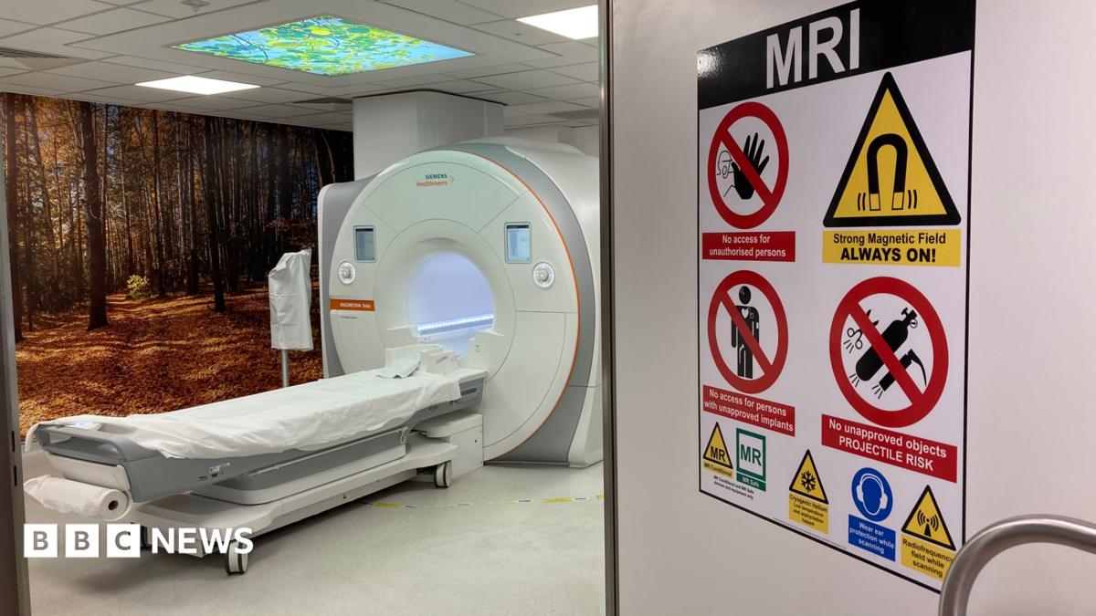 MRI scanner in a hospital room, which has an open door with a warning sign on saying MRI Strong Magnetic Field ALWAYS ON! The machine has a long flat bed that goes into a doughnut like scanner.