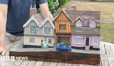 A man poses next to a model of a row of cottages. There are two cottages, one pink and one cream, with a smaller, orange crooked cottage between them. There is a blue car parked outside the orange home. The man has light hair and wears a blue T-shirt and cream shorts. He rests his hands on the table that the model sits on.
