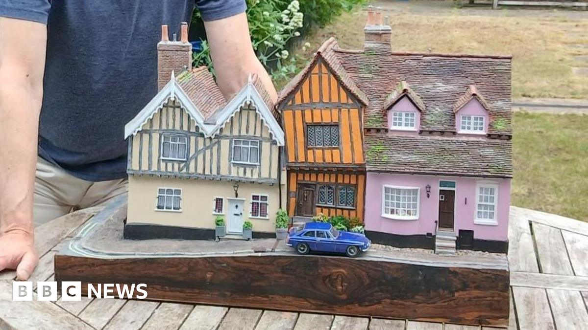 A man poses next to a model of a row of cottages. There are two cottages, one pink and one cream, with a smaller, orange crooked cottage between them. There is a blue car parked outside the orange home. The man has light hair and wears a blue T-shirt and cream shorts. He rests his hands on the table that the model sits on.