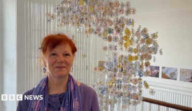 Gillian Taylor, who has red hair and is wearing purple, stands in front of her art installation. The daisies made from the letters hang on fishing wire from the ceiling of the stairwell.