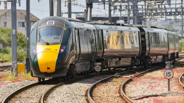 A yellow and dark green GWR branded train leaving a railway station, driving towards the camera.