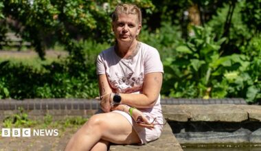 Jackie is sitting with her legs and arms crossed on a concrete wall with greenery behind her. She is wearing a pink T-shirt with a butterfly pattern on the front and pink shorts. Her blonde hair is pulled back in braids. She is wearing a watch and bracelets and is holding a pair of sunglasses in one hand.