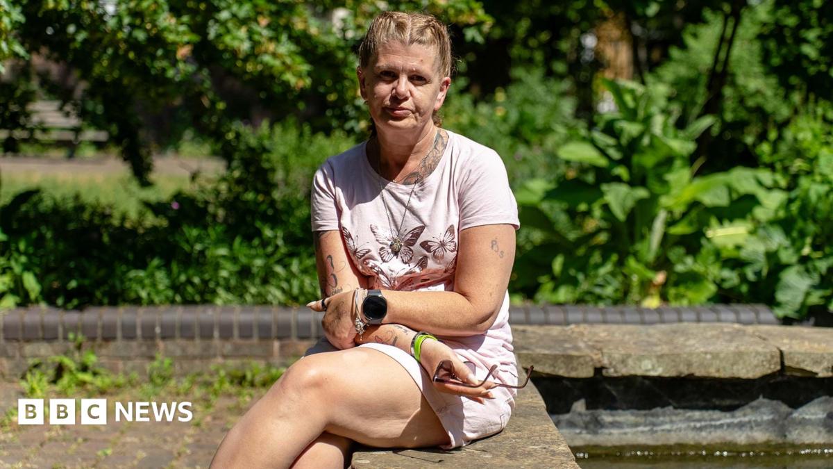 Jackie is sitting with her legs and arms crossed on a concrete wall with greenery behind her. She is wearing a pink T-shirt with a butterfly pattern on the front and pink shorts. Her blonde hair is pulled back in braids. She is wearing a watch and bracelets and is holding a pair of sunglasses in one hand.