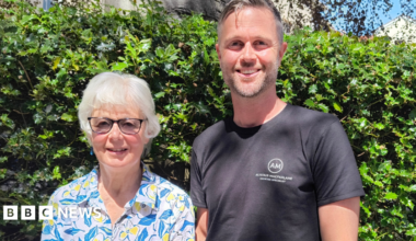 Alastair and his mum Anne are pictured standing in front of a green bush. They are both smiling at the camera. Anne has short white hair and is wearing a white shirt with a blue leaf and yellow flower pattern. She is also wearing black-rimmed glasses. Alastair has short greying hair and stubble. He is wearing a black T-shirt with a white logo.