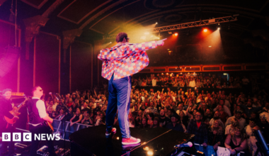 Richard Reeves is seen standing on a piano on a stage facing a crowd of people.