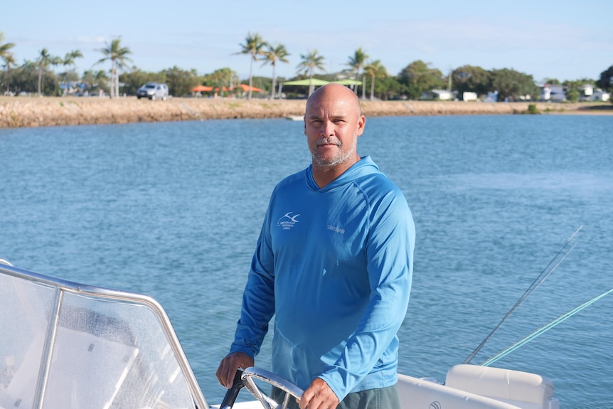 A man in a fishing shirt standing in his boat looking at the camera, with the Bowen foreshore in the background.