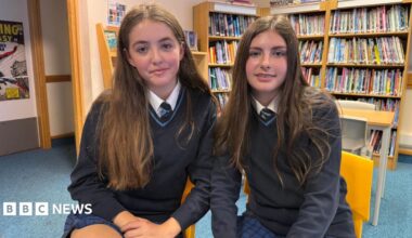 Two pupils at John Cabot Academy, sitting on yellow chairs in the library. They are both wearing a school uniform consisting of a blue and black checked skirt, navy jumper and dark tie. Both have brown hair and are sitting close together.
