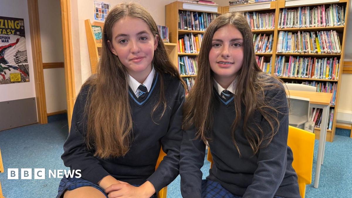 Two pupils at John Cabot Academy, sitting on yellow chairs in the library. They are both wearing a school uniform consisting of a blue and black checked skirt, navy jumper and dark tie. Both have brown hair and are sitting close together.