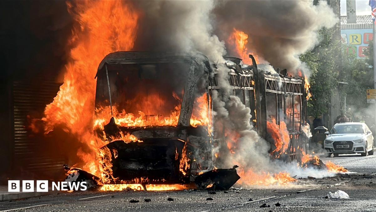A bus is engulfed in flames, with thick black smoke billowing into the air as fire spreads around the vehicle.