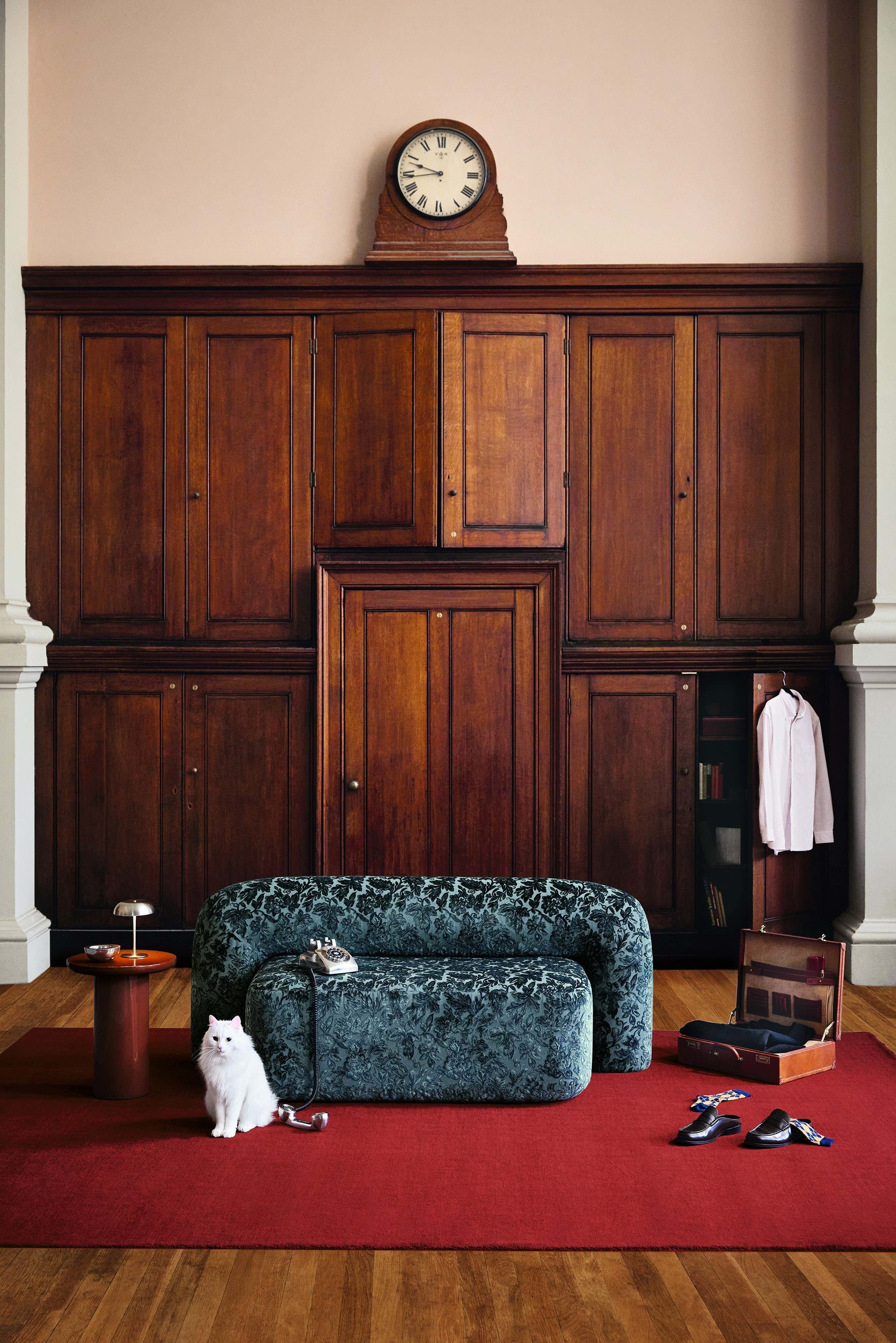 An old, wood-clad room furnished with a chunky love seat in cerulean blue, floral-printed fabric, a red rug, wooden furniture, and scattered clothing.