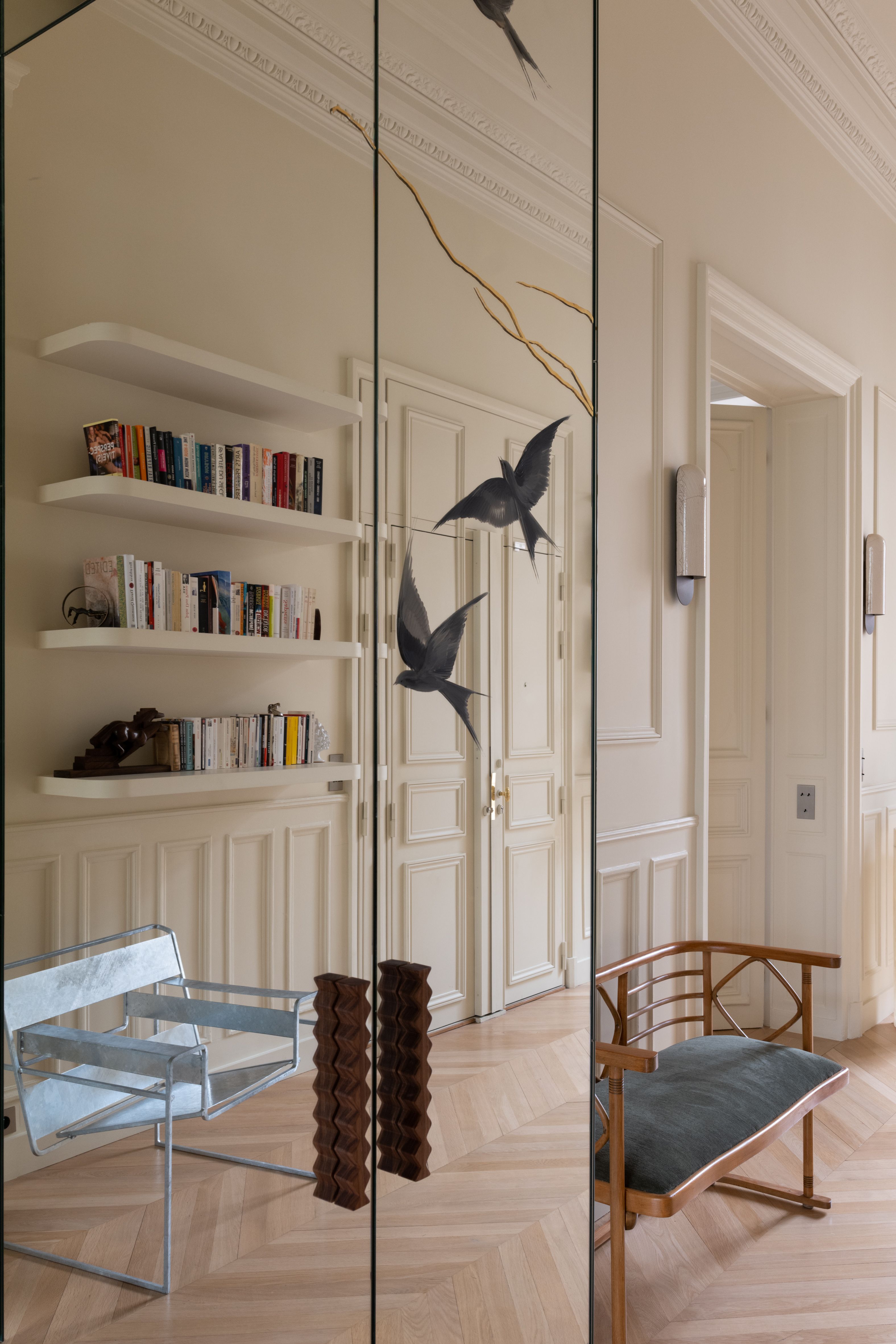 A mirrored closet with two bird stickers on it reflecting the book case at the back of the room in a grand space with off-white walls