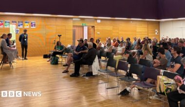 Dozens of people sat inside a school hall for an event. Two people with microphones are sat in front of the audience.
