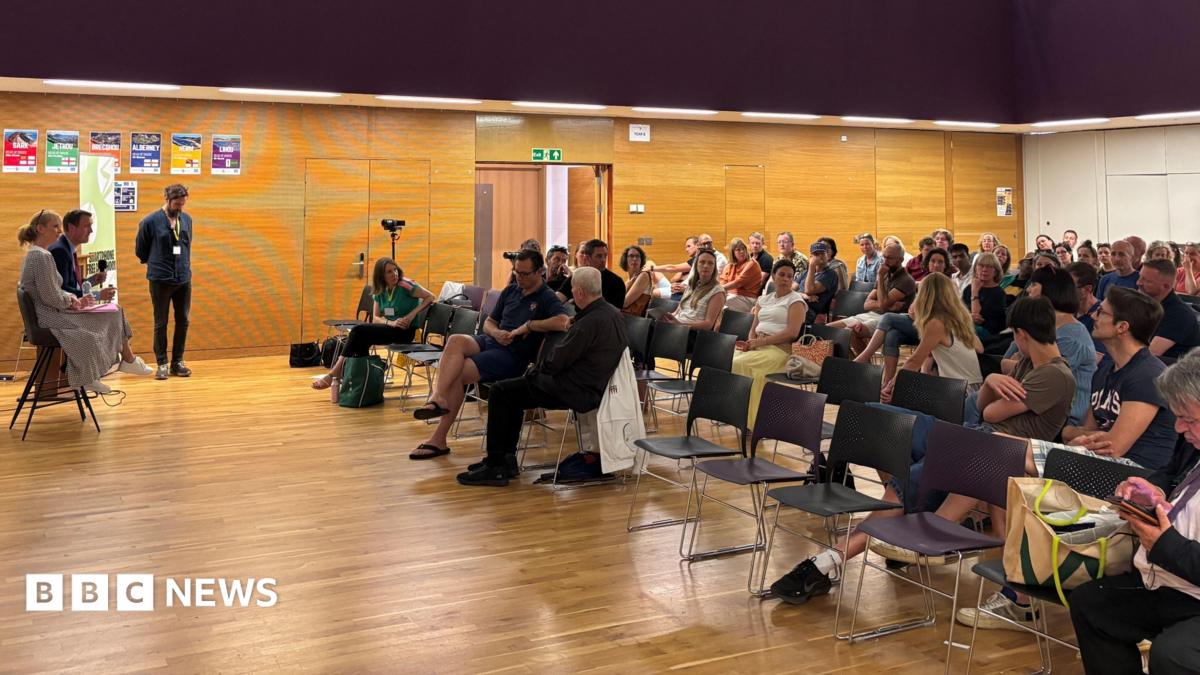 Dozens of people sat inside a school hall for an event. Two people with microphones are sat in front of the audience.