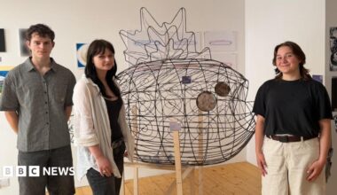 On the left there is a male student wearing a grey shirt and black shorts, a female student wearing a a white overshirt and black jeans, then the metal fish sculpture in the middle with the final female student on the right wearing a black top and white shorts.