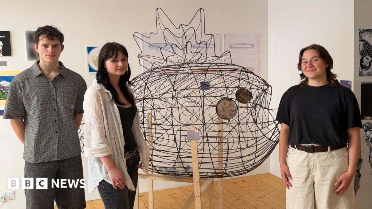 On the left there is a male student wearing a grey shirt and black shorts, a female student wearing a a white overshirt and black jeans, then the metal fish sculpture in the middle with the final female student on the right wearing a black top and white shorts.