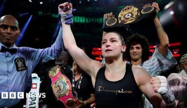 Katie Taylor, centre, with dark tied back hair, wearing a black vest with the word Champion on it. There is a man to her left wearing a blue shirt and a black bow tie, raising her right arm in the air.