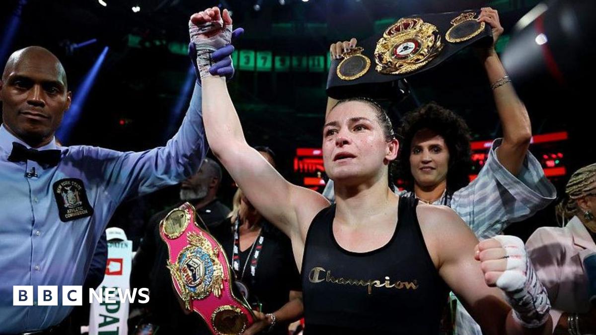 Katie Taylor, centre, with dark tied back hair, wearing a black vest with the word Champion on it. There is a man to her left wearing a blue shirt and a black bow tie, raising her right arm in the air.