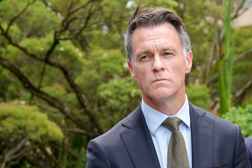 Headshot of serious Chris in a blue suit and tie standing outside, greenery behind.