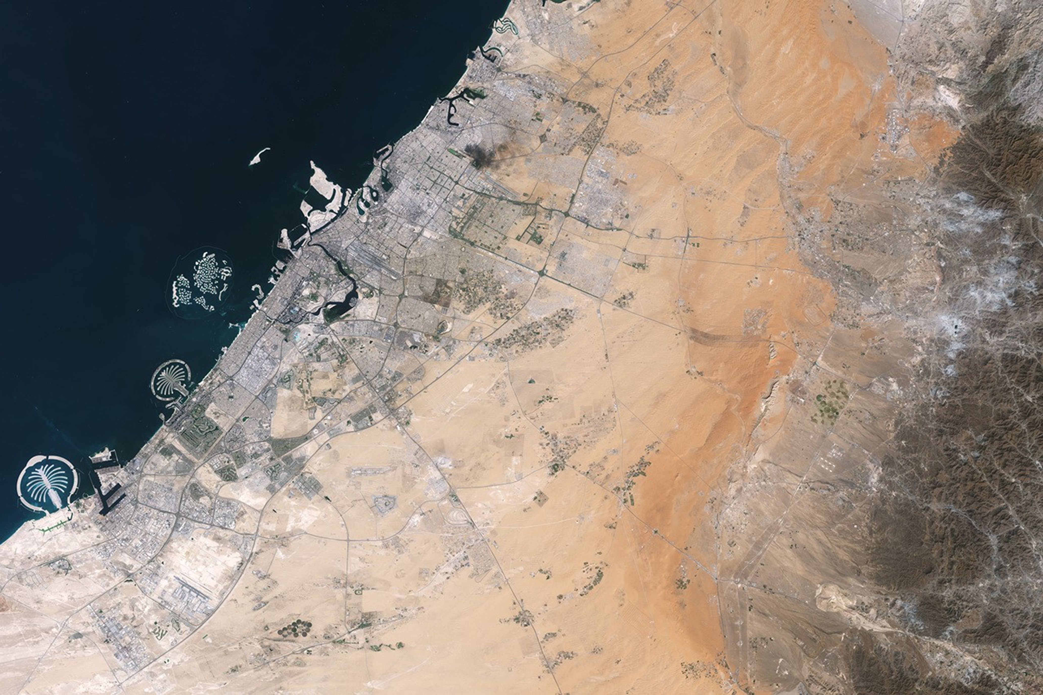 Development associated with Dubai appears as a light gray zone along the coast of the Persian Gulf with the dark gray Al-Hajar Mountains on the far right side of the image. A ridge lies in the middle of the image in a sandy area.