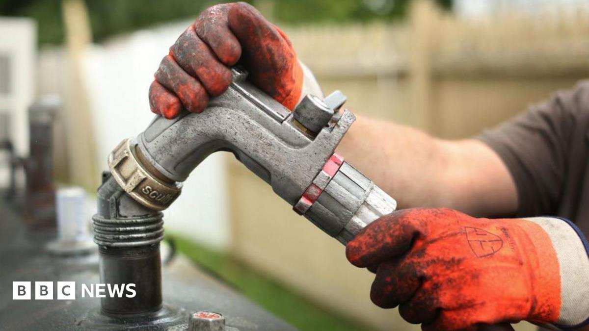 Gloved hands hold the nozzle of an oil hose over what looks like a filling hole of a domestic oil tank. Behind is a small strip of grass and an unpainted wooden fence.