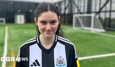 Holly, who is 15, is standing on artificial football turf with a goal, out of focus, in the background. She's wearing a NUFC home shirt and has fair skin and long dark curly hair that's tied in a ponytail. She is smiling.