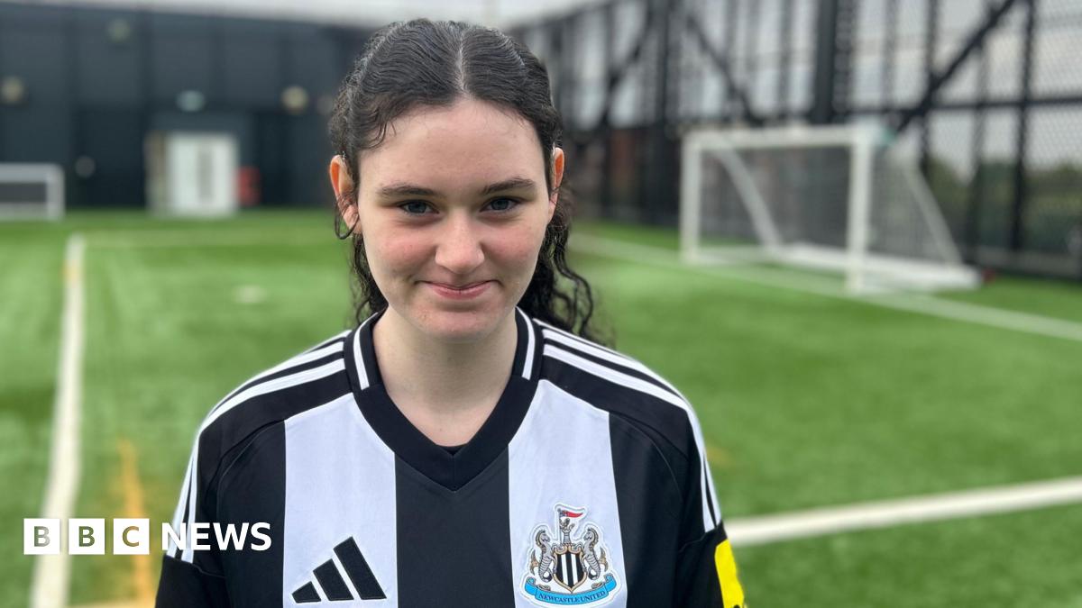 Holly, who is 15, is standing on artificial football turf with a goal, out of focus, in the background. She's wearing a NUFC home shirt and has fair skin and long dark curly hair that's tied in a ponytail. She is smiling.