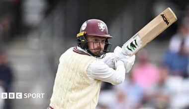 Lewis Goldsworthy in white cable jumper and maroon batting helmet holds his bat up to his right as he plays a shot