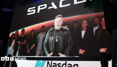 Elon Musk stands at a podium with the Nasdaq logo on it wearing a black t shirt and black leather jacket.