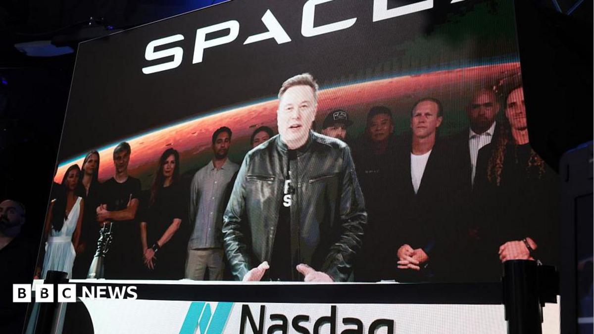 Elon Musk stands at a podium with the Nasdaq logo on it wearing a black t shirt and black leather jacket.
