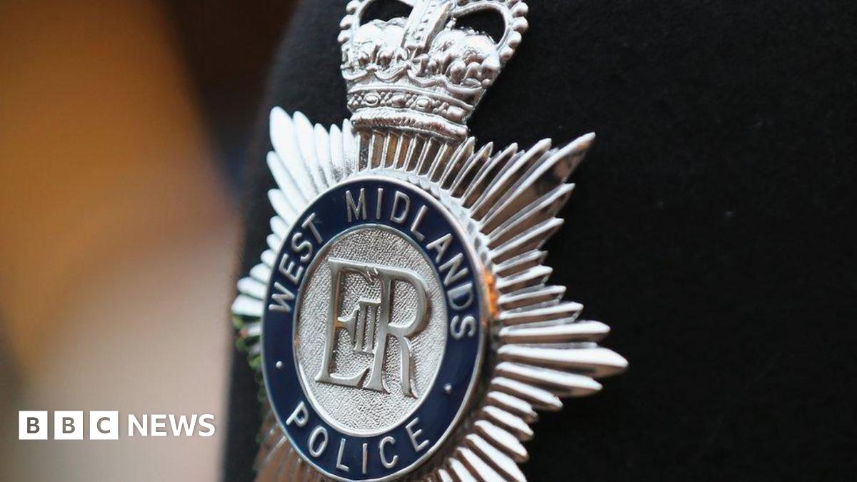 A close-up of a silver police badge on a black jacket, which reads West Midlands Police.