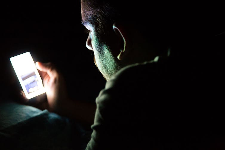 A man, seen from behind, looks at his phone in a dark room.