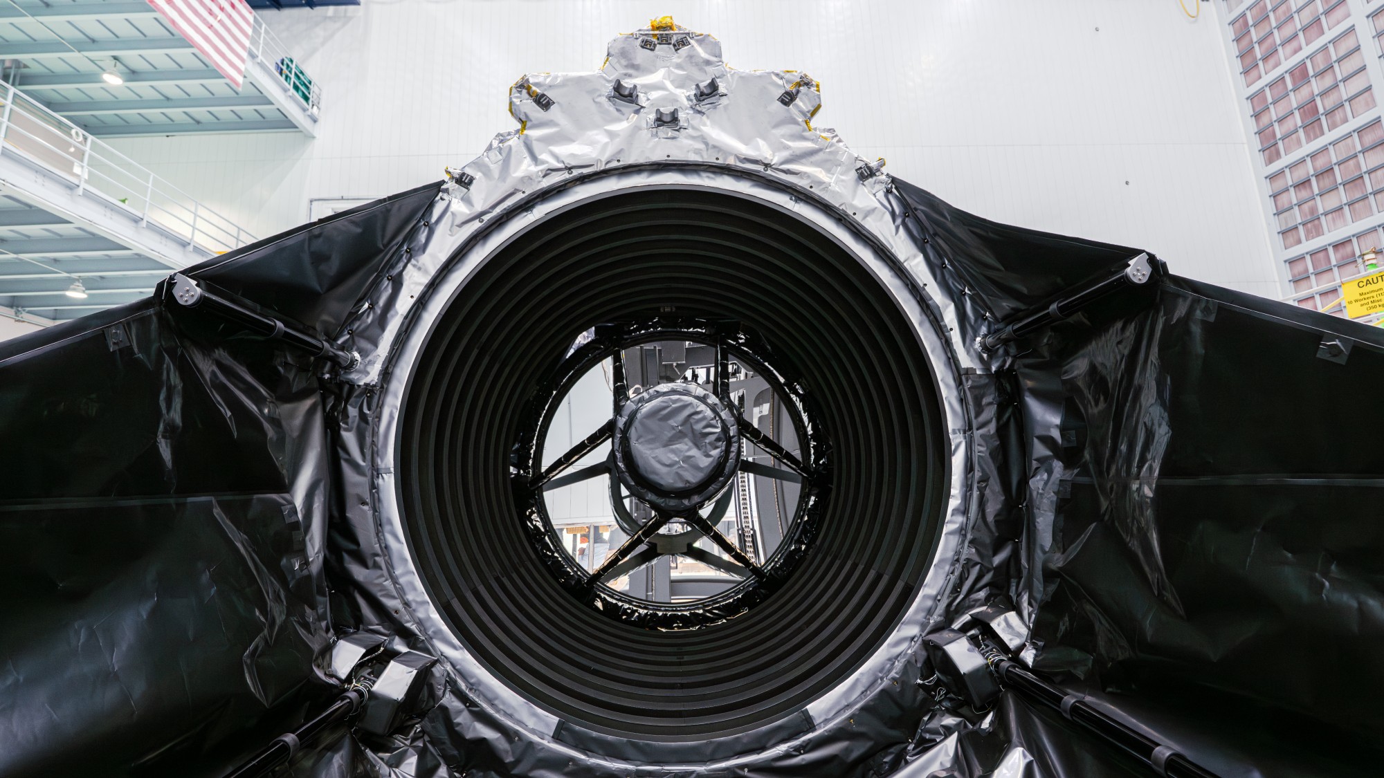technicians in clean suits in a hangar next to a large rectangular spacecraft wrapped in metallic foil