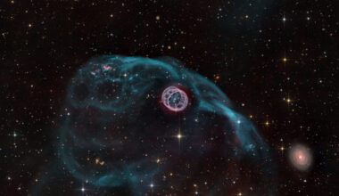 A gaseous structure that resembles a hamster wheel sits near the center of the image. Inside is a bright white dwarf star. There is a larger asymmetric gas bubble surrounding the inner nebula. A galaxy sits to the bottom right. The background is composed of foreground and background stars, as well as distant galaxies.