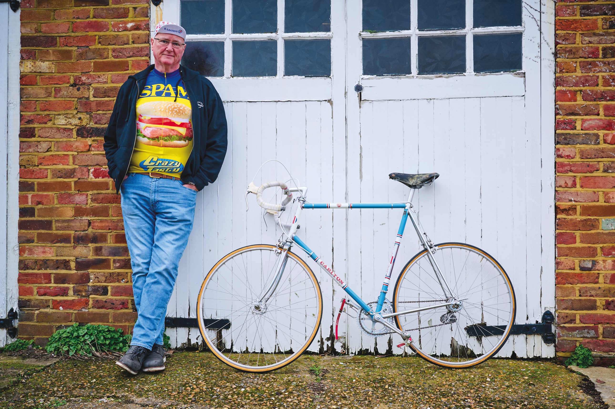 Carlton Kirby standing leaning on his white garage doors alongside his vintage racing bike