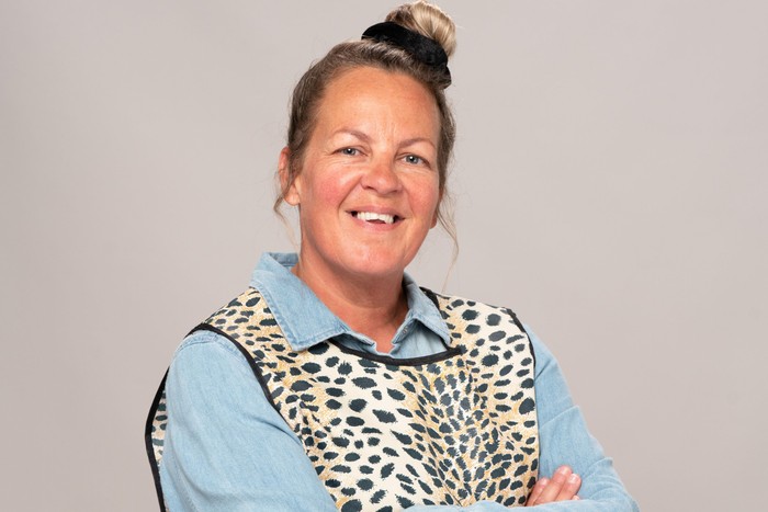 Lorraine Stanley as Karen Taylor. She is wearing a blue shirt and leopard print overall. She is smiling.