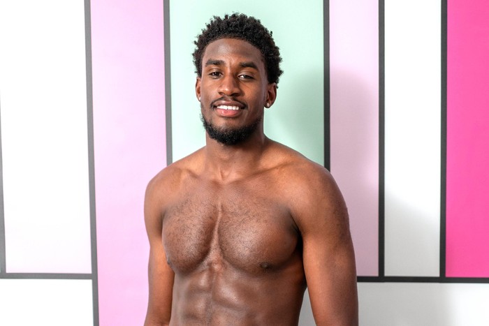 love-island (45) A topless man smiling ahead stood in front of a pastel blue and pink background.