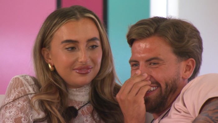 Love Island S13 Yasmin looking at Aidan as they sit next to each other. Aidan is laughing and has one hand covering his mouth.
