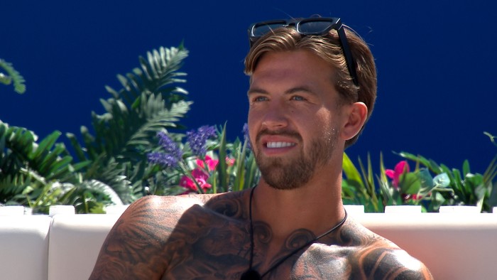 Love Island S13 A topless man with tattoos across his chest, smiling ahead as she sits back at the Love Island fire pit.