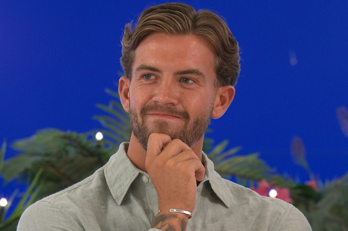 Love Island S13 Aidan smiling ahead with his hand on his chin at the Love Island fire pit.